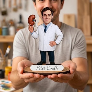 Puede incluir: Una figura plana recortada de un médico con bata blanca, camisa azul y corbata oscura, sosteniendo un modelo anatómico de riñón marrón. La figura se apoya sobre una base circular de madera con una placa blanca que muestra el nombre Peter Smith.