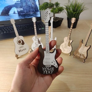 Może przedstawiać: Zestaw sześciu miniaturowych drewnianych gitar, każda z innym imieniem wygrawerowanym na korpusie. Gitary są pomalowane w różnych odcieniach brązu i bieli i są wystawione na drewnianej powierzchni. Gitary są wszystkie w tym samym stylu, z klasycznym kształtem gitary elektrycznej. Tekst na gitarach brzmi "Mario", "Yo", "Brody", "Adolfo" i "YOUR TEXT".