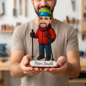 May include: A custom caricature figurine of a skier wearing a red jacket, blue pants, a plaid scarf, and green ski goggles. The figure holds two black ski poles and stands on a wooden base with a white plaque displaying the name Peter Smith.