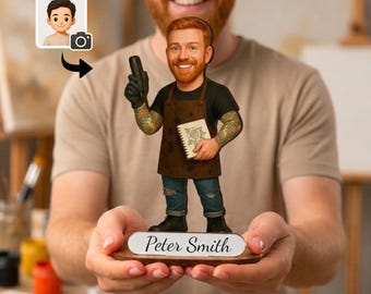 May include: A custom wooden figurine of a tattooed person with a red beard, wearing a black t-shirt, brown apron, and blue jeans. The figure holds a tattoo machine and a sketchbook, standing on a base with the name Peter Smith printed on it.