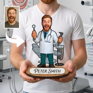 May include: A white t-shirt featuring a cartoon illustration of a male dentist with a red beard, wearing a white lab coat, blue tie, and teal pants. The character holds a dental mirror and tool, standing on a wooden base labeled Peter Smith.
