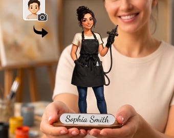 May include: A custom figurine of a person with dark hair in a bun, wearing a white t-shirt, blue jeans, and a black apron with pockets. The figure holds a black airbrush tool and stands on a wooden base with a white plaque reading Sophia Smith.
