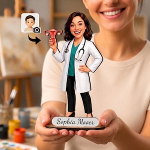 Puede incluir: Una figura cabezona personalizada de un médico con cabello castaño, vistiendo una bata blanca sobre una camisa verde azulado y pantalones negros, sosteniendo un modelo anatómico de útero rosa. La base tiene una placa blanca con el nombre Sophia Moover.