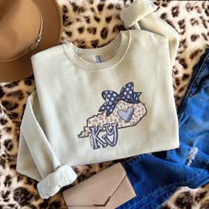 Peut inclure: Un sweat-shirt vert clair avec un motif de l'État du Kentucky, orné d'un imprimé léopard, d'un nœud bleu à pois et d'un cœur bleu. Les lettres "KY" sont en bleu. Le sweat-shirt est associé à un jean bleu et à une pochette beige.