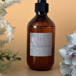 Puede incluir: Una botella de vidrio marrón con un dispensador de bomba negro. La botella está etiquetada con "OLU ESTD 2019" y "SNAIL MUCIN CLEANSER".