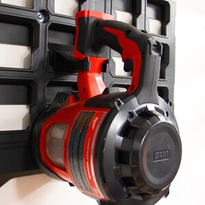 May include: A red and black cordless leaf blower with a black handle and a clear plastic dust container. The blower is mounted on a black wall organizer.