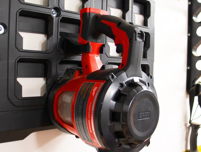 May include: A red and black cordless leaf blower with a black handle and a clear plastic dust container. The blower is mounted on a black wall organizer.
