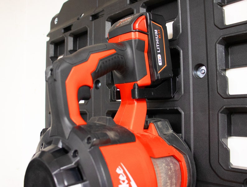 May include: A red and black cordless leaf blower with a black and orange battery attached to the side. The battery is labeled "18V LITHIUM-ION". The blower is mounted on a black wall-mounted storage system.