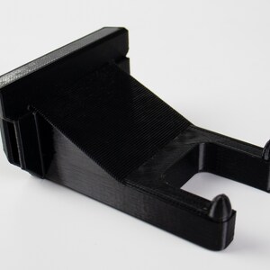 May include: Black plastic wall mount with two small protrusions on the bottom. The mount is designed to hold a small object.