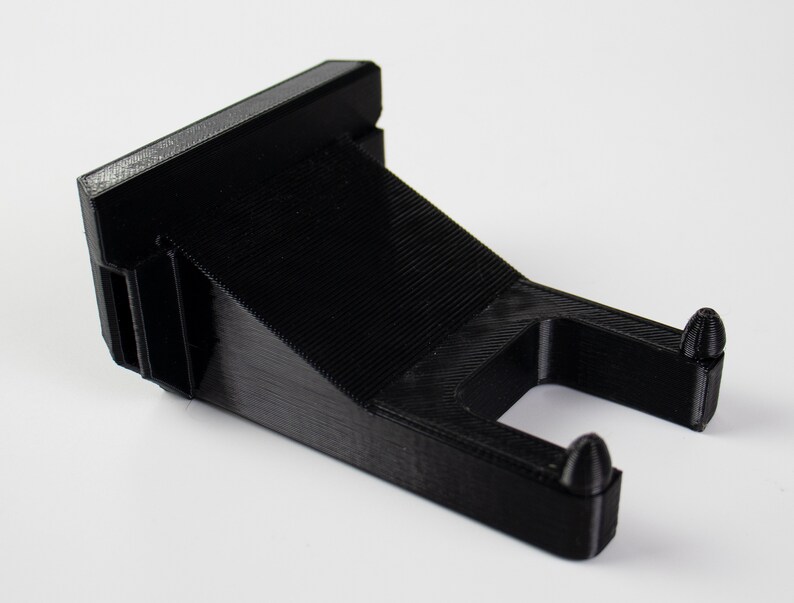 May include: Black plastic wall mount with two small protrusions on the bottom. The mount is designed to hold a small object.