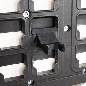May include: A black plastic wall-mounted organizer with multiple slots for holding various items. The organizer has a small black plastic hook on the right side.