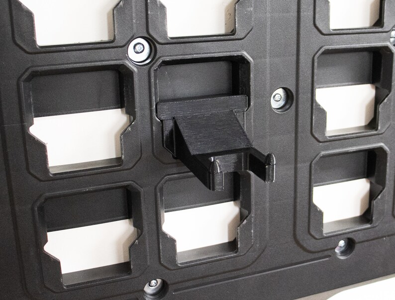 May include: A black plastic wall-mounted organizer with multiple slots for holding various items. The organizer has a small black plastic hook on the right side.
