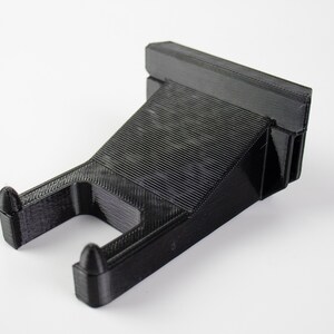 May include: A black plastic wall mount with a hook for hanging items. The mount has a textured surface and a rectangular shape.