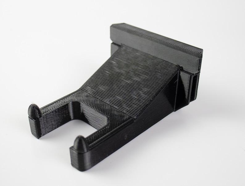 May include: A black plastic wall mount with a hook for hanging items. The mount has a textured surface and a rectangular shape.