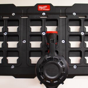 May include: Black Milwaukee Packout wall-mounted storage system with 18 compartments and two tools hanging on the front. The tools are a black and red power drill and a black circular saw.