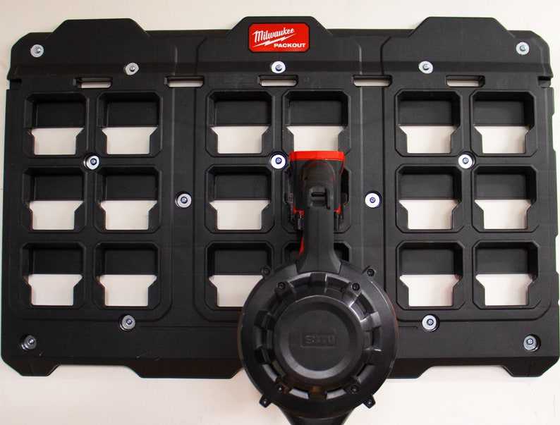 May include: Black Milwaukee Packout wall-mounted storage system with 18 compartments and two tools hanging on the front. The tools are a black and red power drill and a black circular saw.