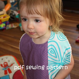 Puede incluir: Un niño pequeño con un babero morado y turquesa con lunares blancos y un patrón de espiral. El babero tiene el texto "pdf sewing pattern" escrito en él. El niño está sosteniendo un puñado de uvas.