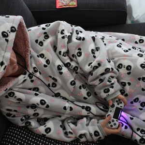 Könnte beinhalten: Eine Person trägt eine graue und rosafarbene Kapuzendecke mit Panda-Druck. Die Decke ist überdimensioniert und hat eine Tasche auf der Vorderseite. Die Person hält einen Videospiel-Controller.