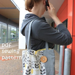 May include: A gray tote bag with a white and yellow floral pattern and a large brown button. The bag has a long strap and is being carried by a person.