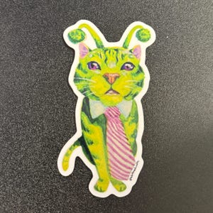 May include: A die-cut sticker featuring a lime green cat with purple eyes, wearing a white collared shirt and a pink and white striped necktie. The cat has two green antennae with fuzzy tips extending from its head and a white border outline.