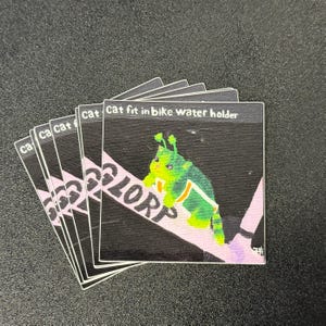 Pode incluir: Uma pilha de autocolantes quadrados com um gato de desenho animado em tons de verde e amarelo, com o texto "Cat fit in bike water holder". Os autocolantes têm um fundo preto com um design rosa e branco.