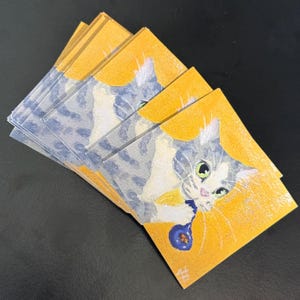 May include: A stack of rectangular stickers featuring a painting of a grey and white tabby cat with green eyes on a bright yellow background. The cat is holding a small blue object with its paws. Each sticker has a glossy finish and clear edges.