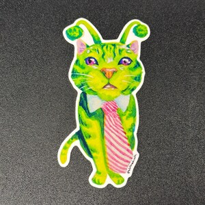 Pode incluir: Um autocolante extravagante com um gato verde de olhos roxos, usando uma camisa branca com gola e uma gravata rosa listrada. O gato tem antenas únicas em forma de folha. O autocolante está em um fundo escuro.