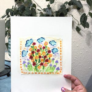May include: A colorful floral embroidery on a white background. The artwork features a vibrant bouquet of red, orange, and yellow flowers, with blue cloud accents and purple and green foliage. The piece is held by a hand with purple nail polish.