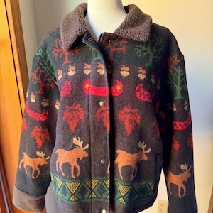 May include: A dark brown jacket featuring a brown sherpa collar and cuffs. The fabric displays a repeating pattern of orange moose, red maple leaves, green trees, and red canoes, with a geometric yellow and green border at the hem.