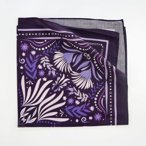 May include: Purple bandana with a floral pattern and a white border. The bandana has a white and purple floral design with a repeating pattern. The bandana is folded in half and the text "VOILE" is visible on the fold.
