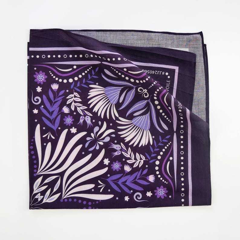 May include: Purple bandana with a floral pattern and a white border. The bandana has a white and purple floral design with a repeating pattern. The bandana is folded in half and the text "VOILE" is visible on the fold.
