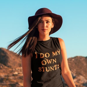 May include: A woman wearing a black sleeveless shirt with gold lettering that says "I do my own stunts" and a brown wide-brimmed hat. She is standing in front of a mountain range.