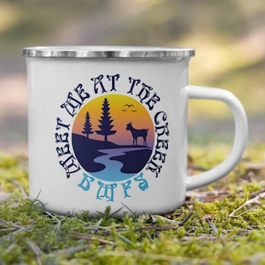Puede incluir: Taza de camping de esmalte blanco con un gráfico de puesta de sol azul, amarillo y naranja. El gráfico incluye un río, dos árboles y una cabra. El texto en la taza dice "Meet me at the Green Bluffs".