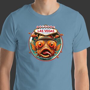 May include: A light blue t-shirt with a graphic of a cartoon fish wearing sunglasses and a hat. The fish is in front of a sign that says "Welcome to Fabulous Las Vegas, Nevada".
