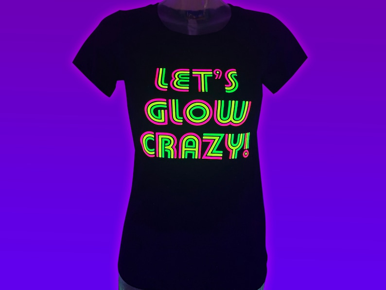 Puede incluir: Camiseta negra con el texto "LET'S GLOW CRAZY!" en verde ne&oacute;n, rosa y amarillo. El texto est&aacute; centrado en la parte delantera de la camiseta. La camiseta tiene cuello redondo y mangas cortas.
