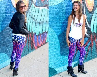May include: Two women wearing colourful leggings with a pattern of circles. The leggings are a gradient of blue, purple, pink and yellow. The women are wearing black boots and are standing in front of a blue and white brick wall.