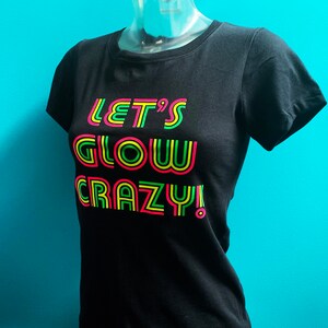 Puede incluir: Camiseta negra con el texto "LET'S GLOW CRAZY!" en rosa ne&oacute;n, amarillo y verde. La camiseta se muestra en un maniqu&iacute; sobre un fondo turquesa. El texto est&aacute; delineado en un color ne&oacute;n.
