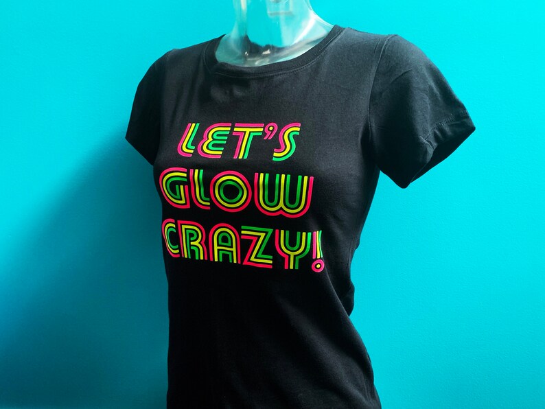 Puede incluir: Camiseta negra con el texto "LET'S GLOW CRAZY!" en rosa ne&oacute;n, amarillo y verde. La camiseta se muestra en un maniqu&iacute; sobre un fondo turquesa. El texto est&aacute; delineado en un color ne&oacute;n.