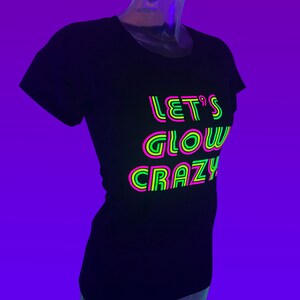 Puede incluir: Camiseta negra con el texto de ne&oacute;n "LET'S GLOW CRAZY" en rosa, verde y amarillo. La camiseta se muestra en un maniqu&iacute; sobre un fondo p&uacute;rpura. El texto est&aacute; superpuesto con un contorno de ne&oacute;n.