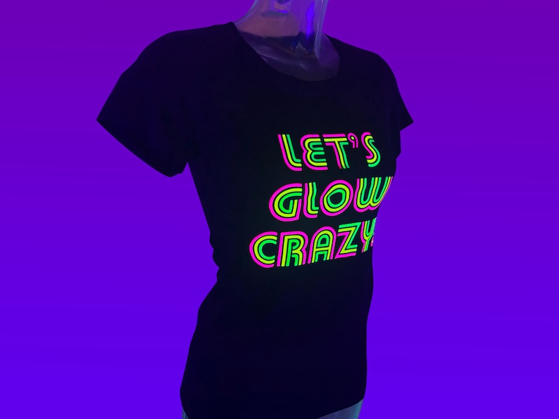Puede incluir: Camiseta negra con el texto de ne&oacute;n "LET'S GLOW CRAZY" en rosa, verde y amarillo. La camiseta se muestra en un maniqu&iacute; sobre un fondo p&uacute;rpura. El texto est&aacute; superpuesto con un contorno de ne&oacute;n.
