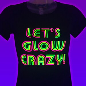 Puede incluir: Camiseta negra con el texto ne&oacute;n "LET'S GLOW CRAZY!" en verde, rosa y amarillo. La camiseta es de cuello redondo y se muestra en un maniqu&iacute;. El fondo es un degradado de color p&uacute;rpura.