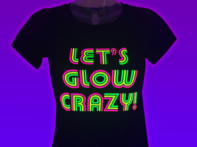 Puede incluir: Camiseta negra con el texto ne&oacute;n "LET'S GLOW CRAZY!" en verde, rosa y amarillo. La camiseta es de cuello redondo y se muestra en un maniqu&iacute;. El fondo es un degradado de color p&uacute;rpura.