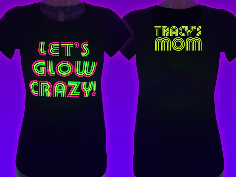 Puede incluir: Dos camisetas negras con texto de ne&oacute;n. Una dice "LET'S GLOW CRAZY!" en verde, rosa y amarillo. La otra dice "TRACY'S MOM" en amarillo. Ambas camisetas tienen cuello redondo.