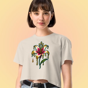 Puede incluir: Un top corto color crema con un diseño floral vibrante. El gráfico representa una flor estilizada con pétalos rosas y blancos, delineados en negro, con acentos dorados goteando. Una hoja y un tallo verdes completan el diseño.