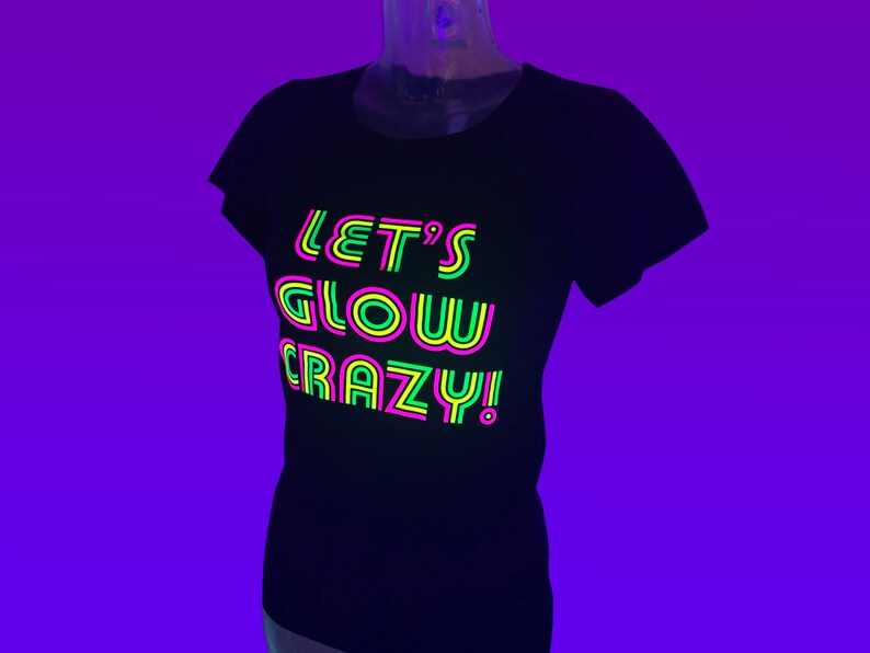 Puede incluir: Camiseta negra con el texto de ne&oacute;n "LET'S GLOW CRAZY!" en rosa, verde y amarillo. La camiseta se muestra en un maniqu&iacute; sobre un fondo p&uacute;rpura. El texto est&aacute; dise&ntilde;ado para brillar bajo luz negra.