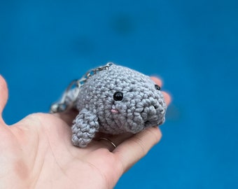 May include: A small gray crocheted manatee keychain with black eyes and pink cheeks. The keychain is held in a hand against a blue background.