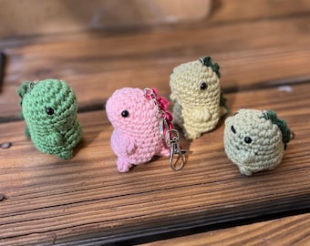 May include: Four crocheted dinosaur keychains in green, pink, and yellow. The dinosaurs have small black eyes and are attached to silver keychains. The keychains are arranged on a wooden surface.