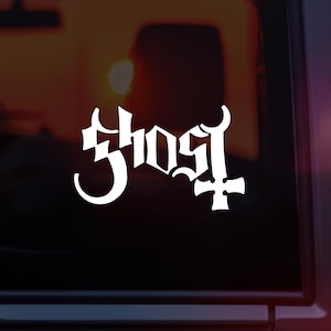 May include: White vinyl decal of the word "Ghost" in a stylized font with a cross underneath. The decal is on a dark red car window.