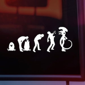 May include: A white vinyl sticker depicting the evolution of a human into an alien. The sticker shows a series of figures, starting with a single-celled organism, then a human in various stages of development, and finally an alien with a large head and a long tail.