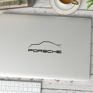 May include: A silver laptop computer with a black outline of a Porsche car and the word "PORSCHE" in black text.